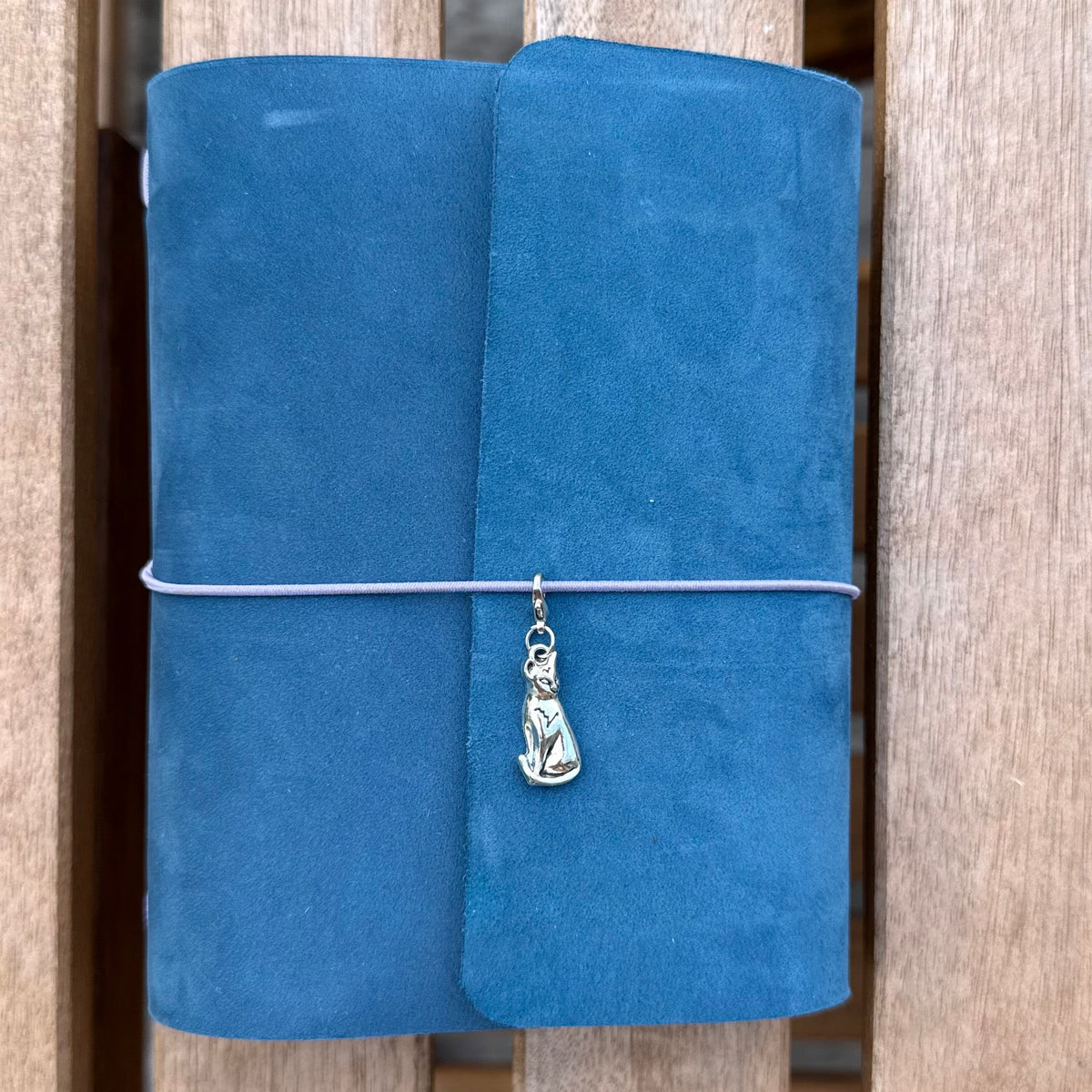 A rolled blue journal or notebook wrapped with a thin elastic band and adorned with a small silver pendant, displayed against a wooden slatted background.