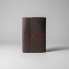 A dark brown leather journal with a red elastic closure cord and a small metal charm, resting on a wooden slatted surface.