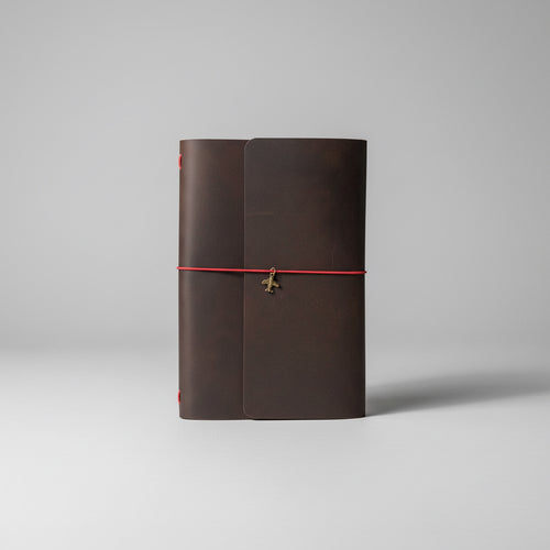 A dark brown leather journal with a red elastic closure cord and a small metal charm, resting on a wooden slatted surface.